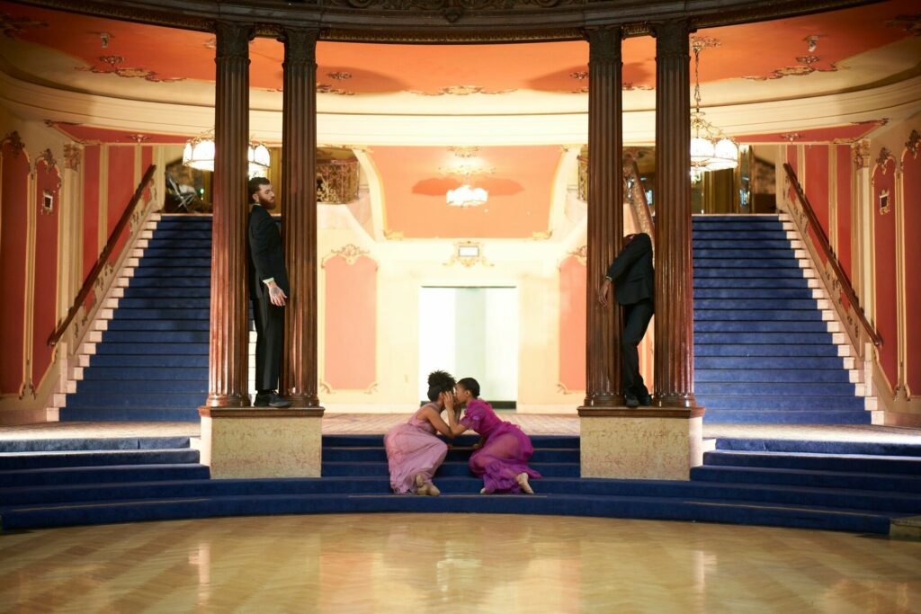 Two women sitting on a staircase with their faces touch. Two men stand behind pillars of the staircase.